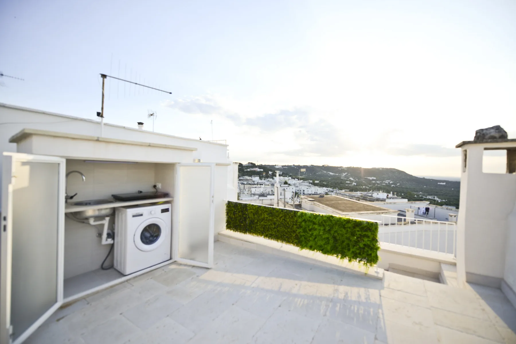Una terraza en la azotea con una lavadora en un rincón de servicio exterior y vistas lejanas del paisaje.