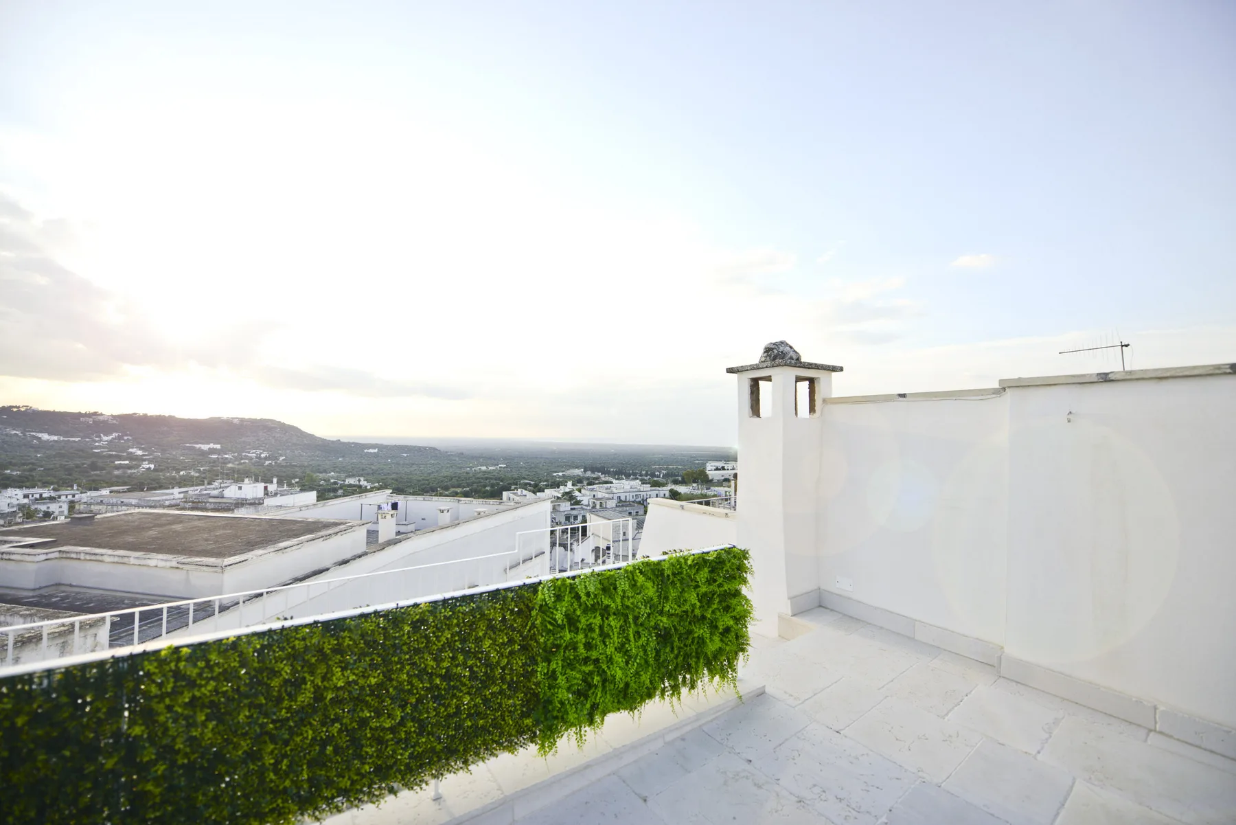 Una terraza en la azotea con una barandilla cubierta de plantas da a edificios y colinas lejanas.