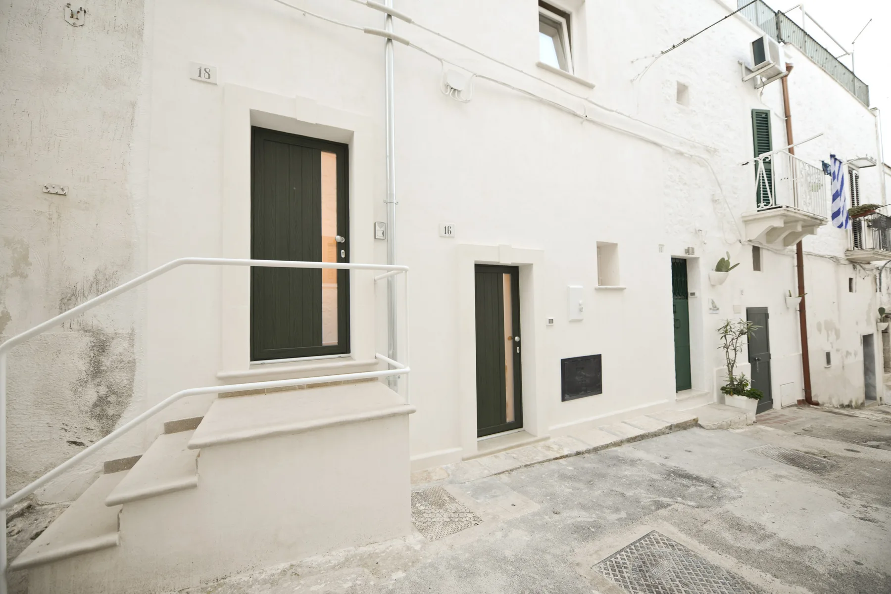 Fachadas blancas con puertas oscuras y una pequeña escalera exterior junto a una calle pavimentada.