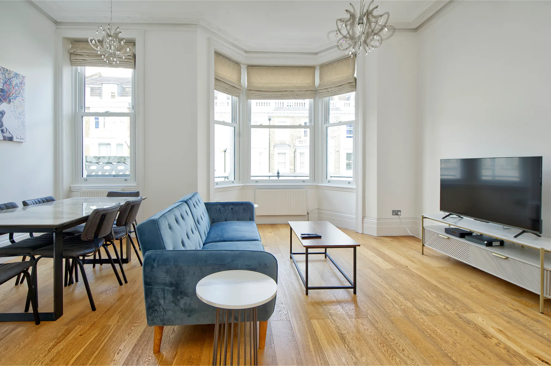 A bright living and dining room with a blue sofa, large windows, and a television.