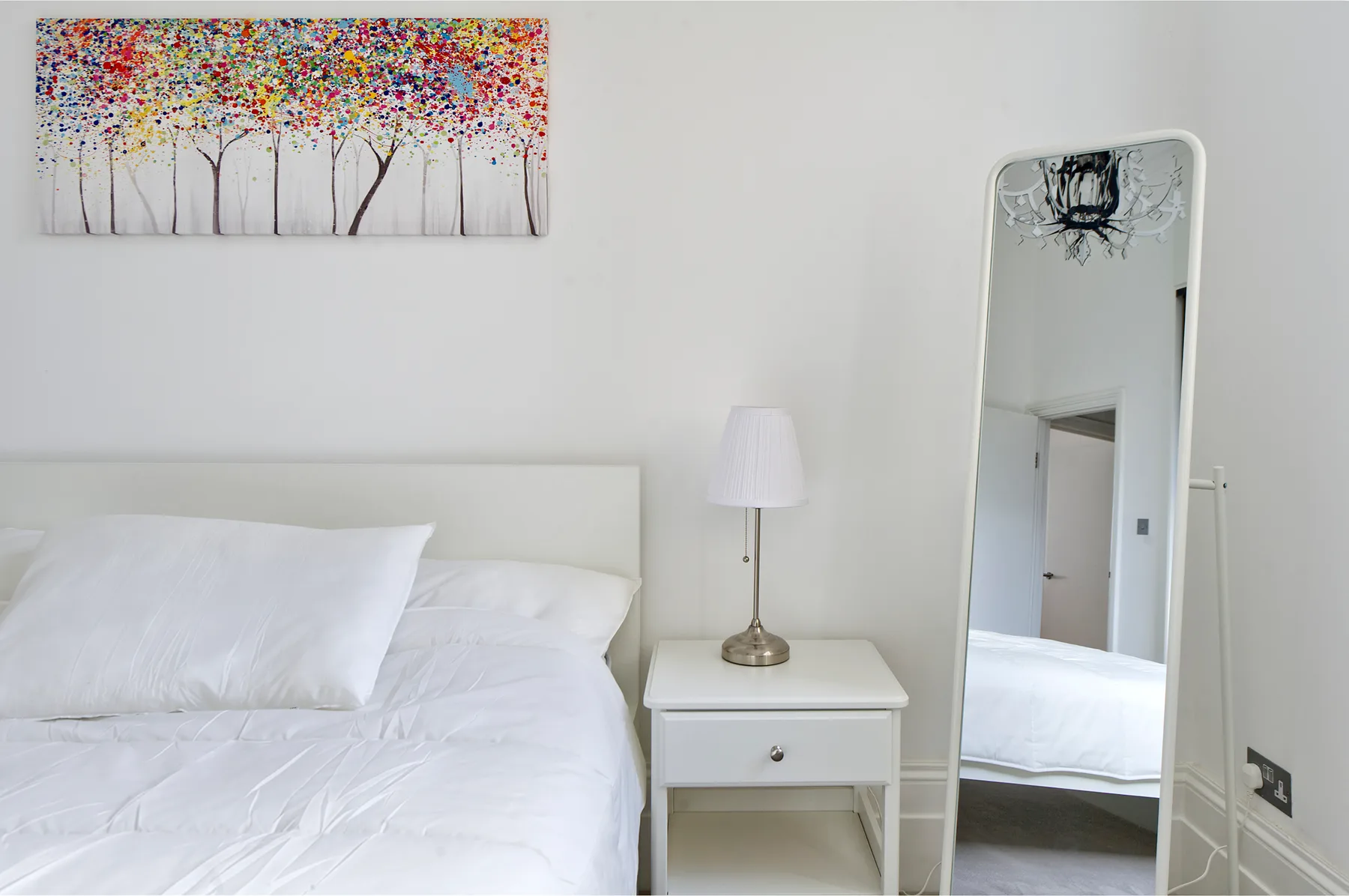 A bright bedroom with white bedding, a bedside table, and colorful tree artwork.
