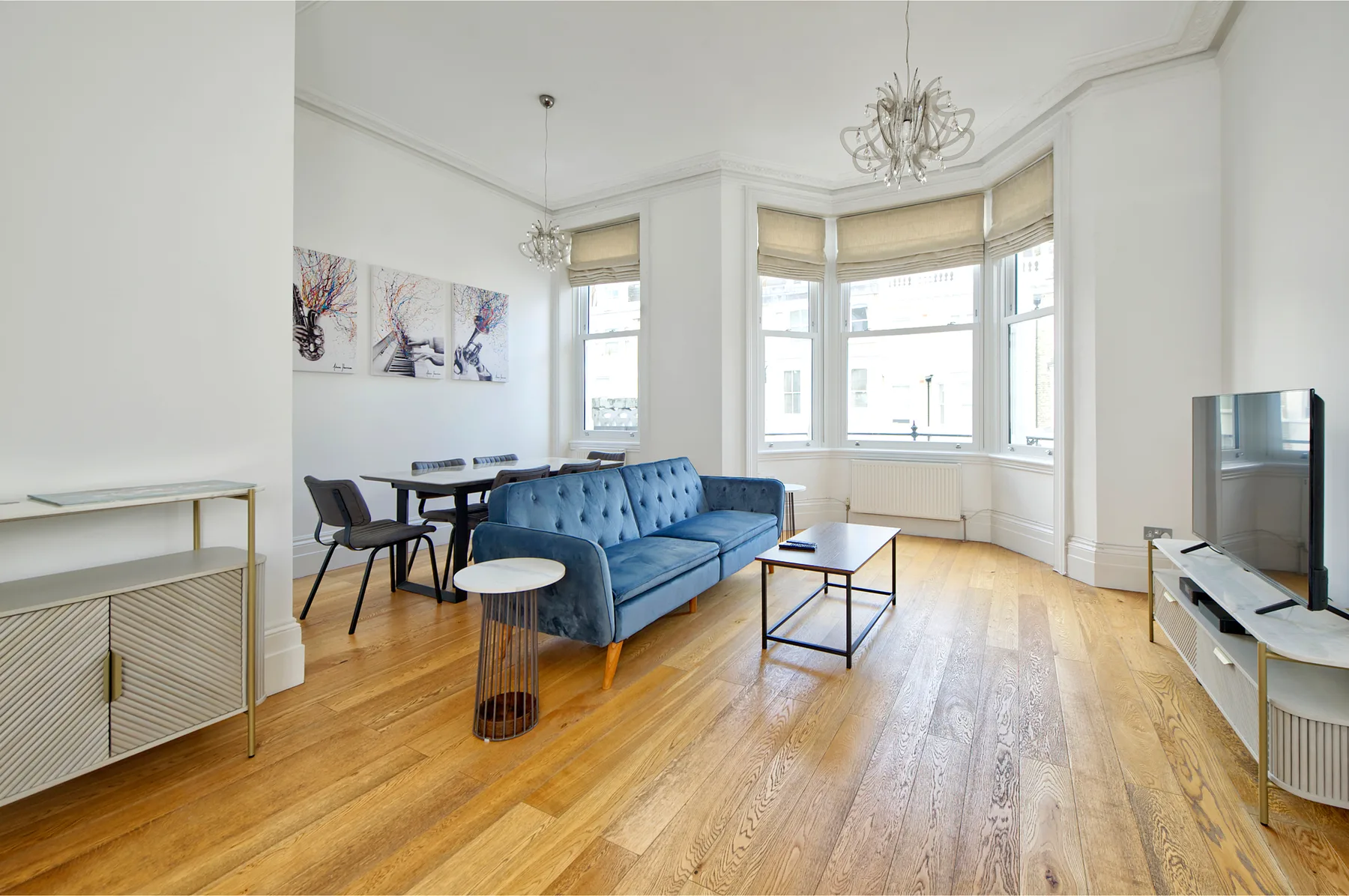 A bright living room with blue seating, a dining area, and large bay windows.