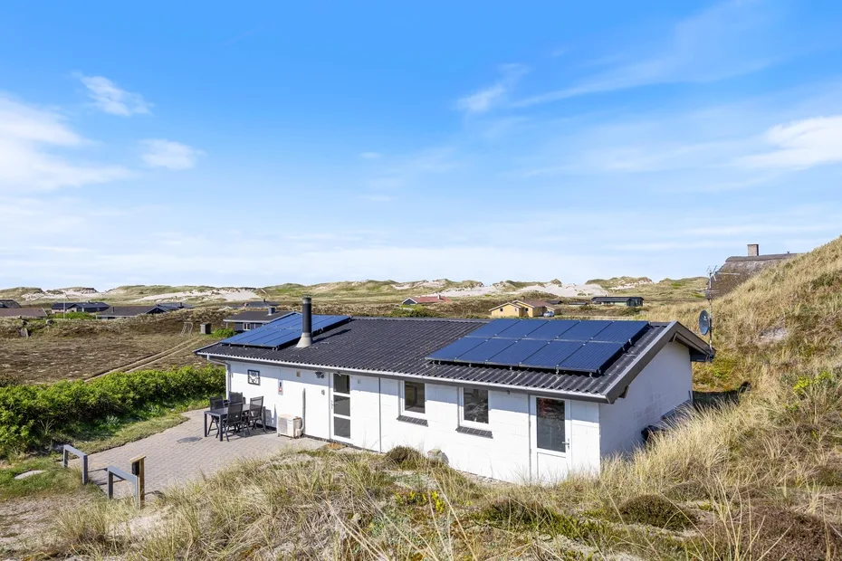 Una casa blanca con paneles solares en el techo se encuentra entre dunas cubiertas de hierba bajo un cielo azul.