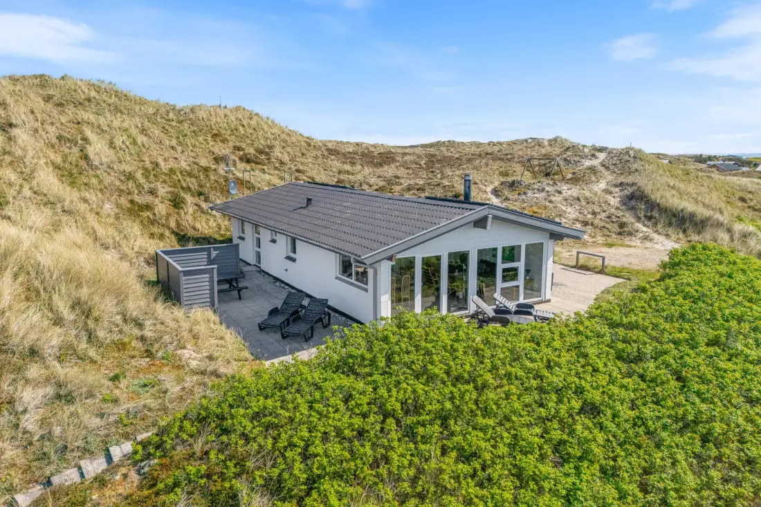 Una casa moderna está entre dunas cubiertas de hierba, con un patio junto a grandes ventanas.