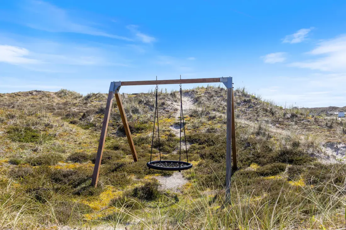 Un columpio cuelga de una estructura de madera entre dunas cubiertas de hierba, bajo un cielo azul.