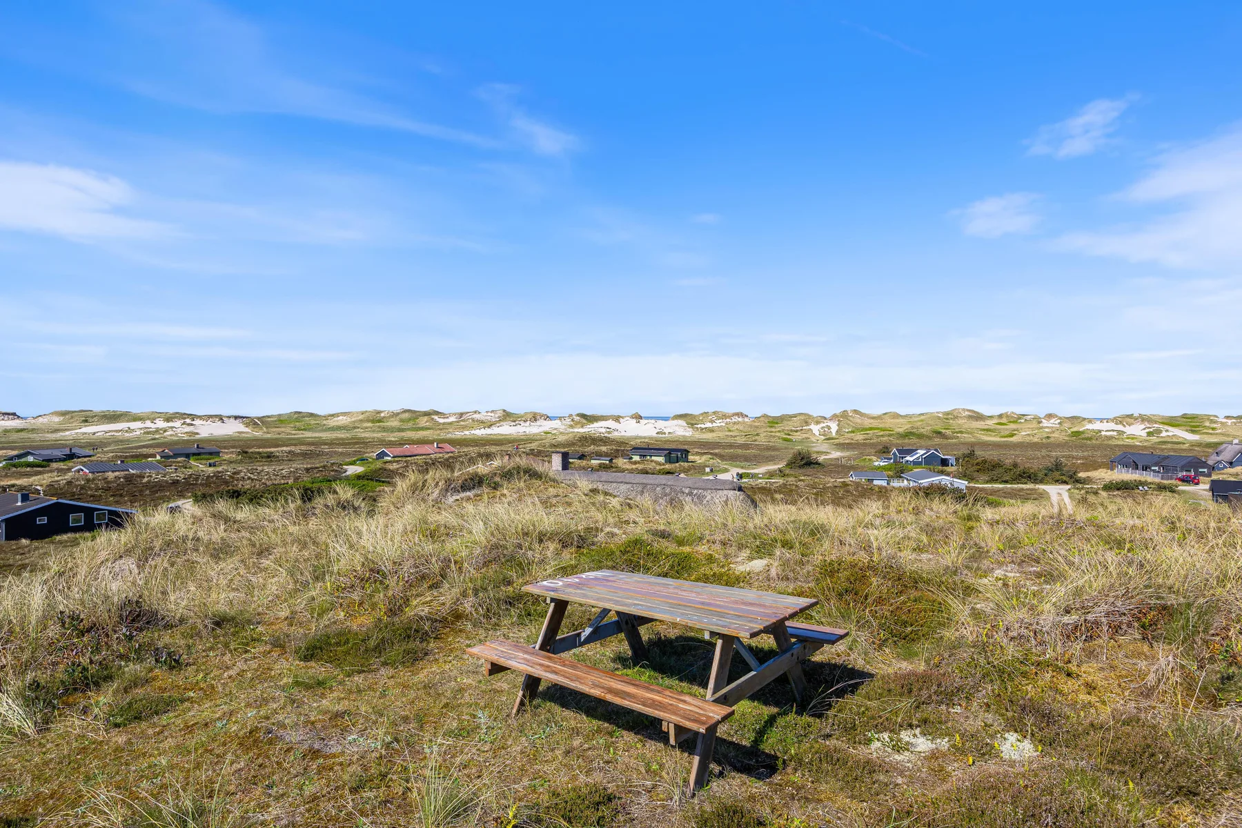 Una mesa de picnic está entre dunas cubiertas de hierba, con casas dispersas a lo lejos.