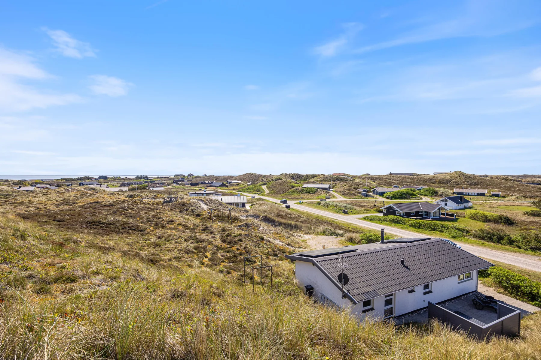 Una casa está entre dunas cubiertas de hierba, junto a una carretera bajo el cielo azul.