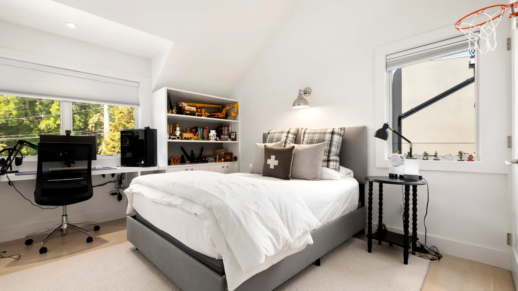 A bright bedroom with a bed, desk, and basketball hoop by the window.