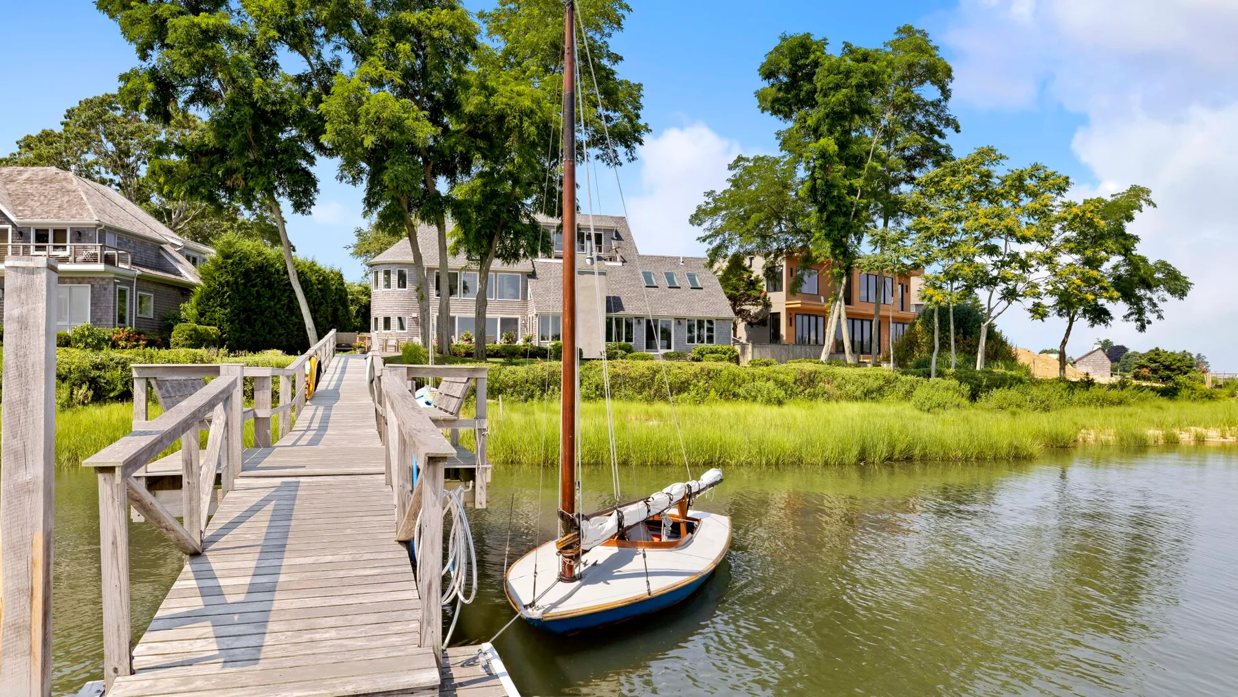 A wooden dock and sailboat sit beside a grassy waterfront with houses and trees.