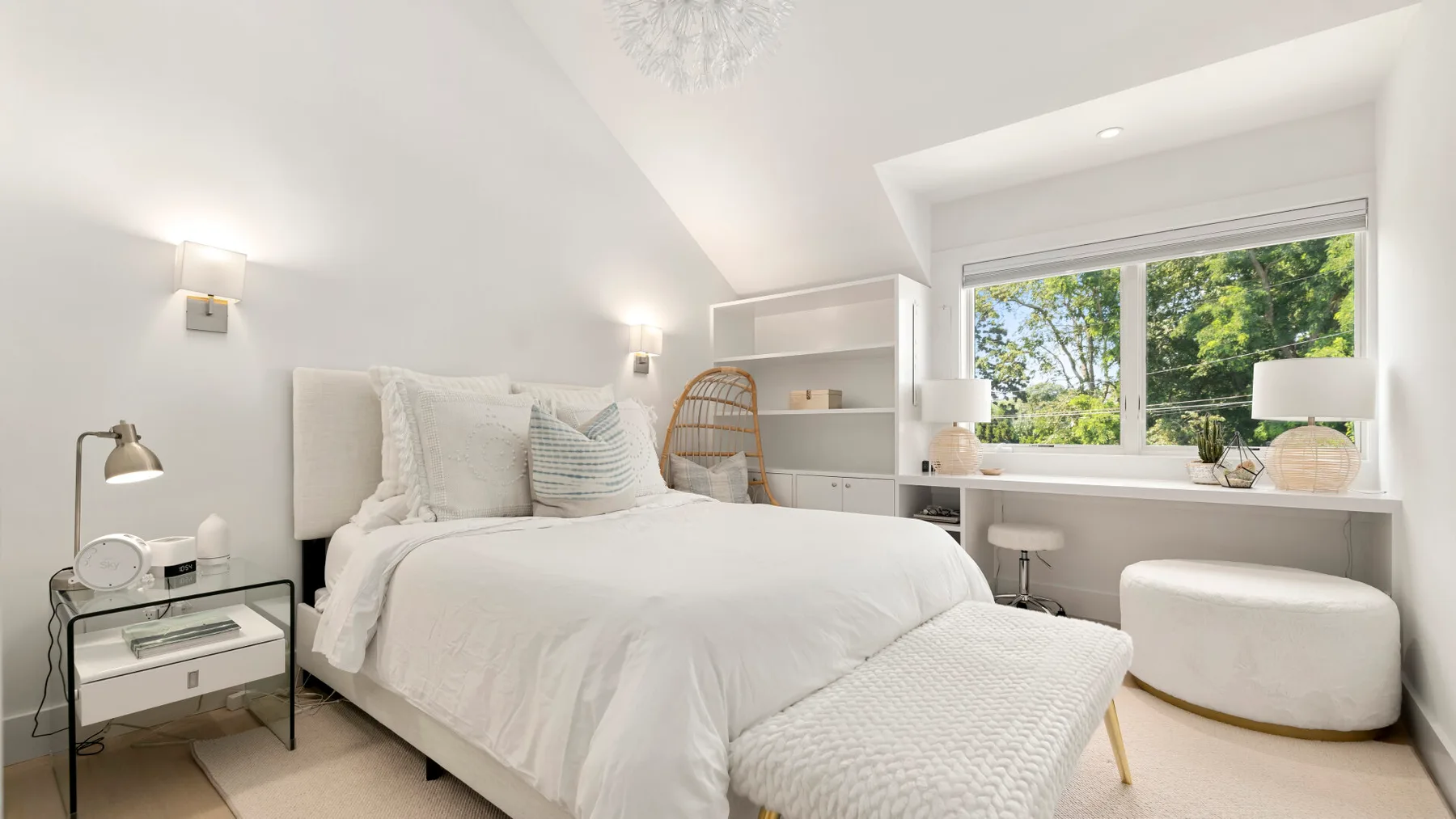 A bright bedroom features a white bed beside a large window overlooking trees.