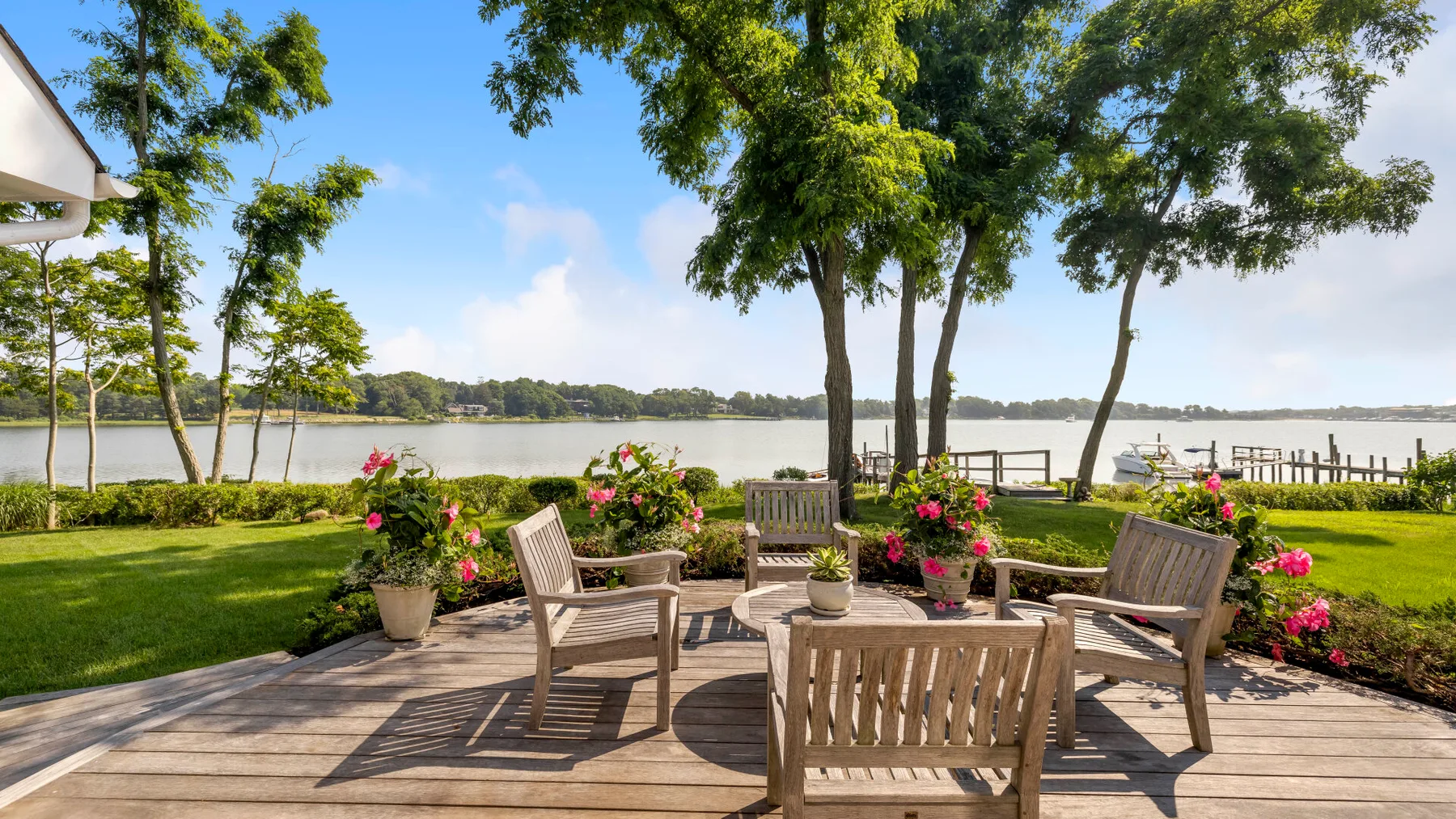 A lakeside patio with wooden seating, flowering plants, and shade trees.