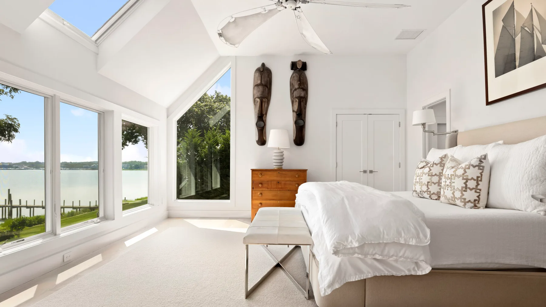 A bright bedroom with large windows and a bed dressed in white linens.