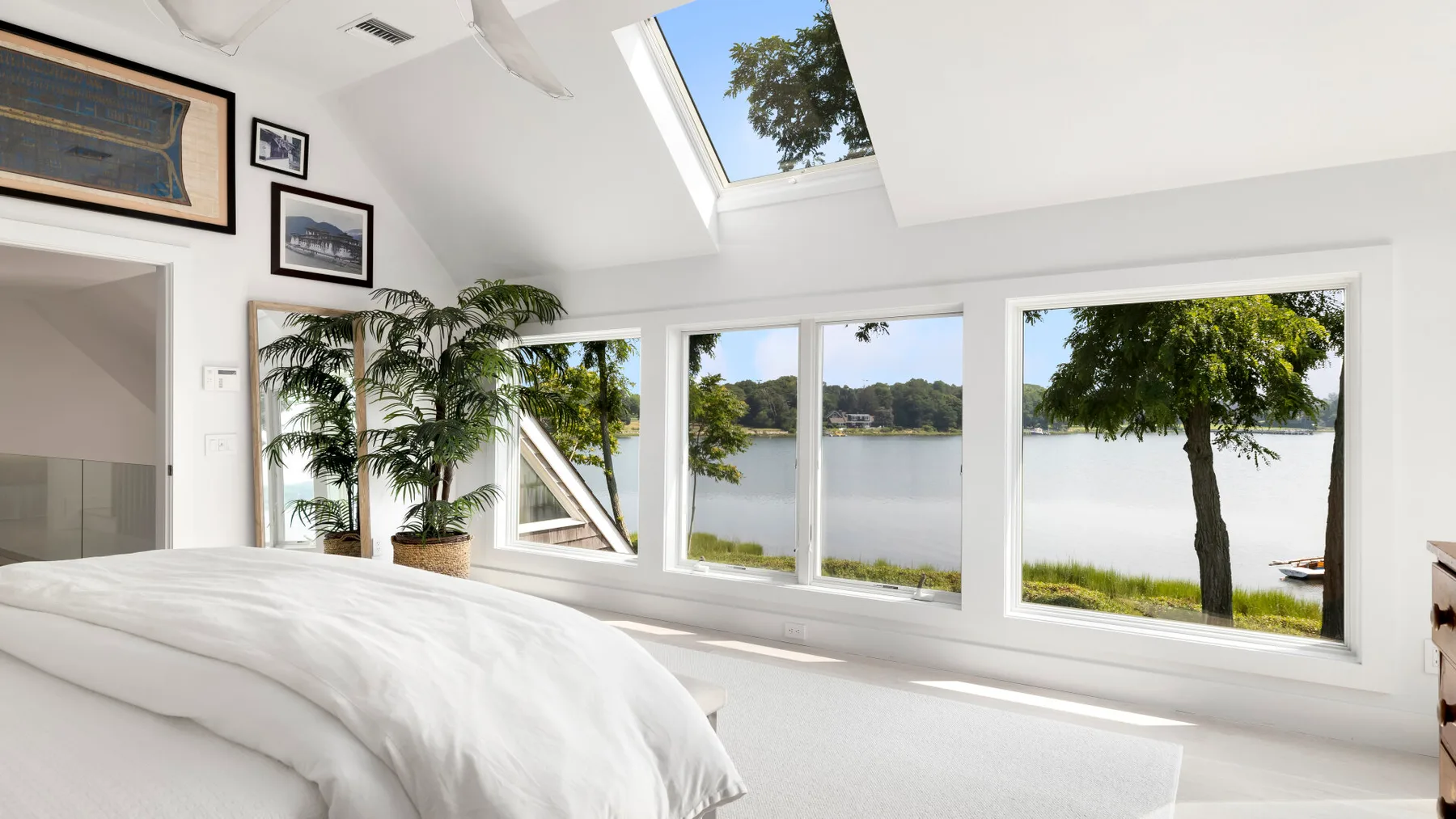 A bright bedroom has wide windows overlooking a lake and trees.
