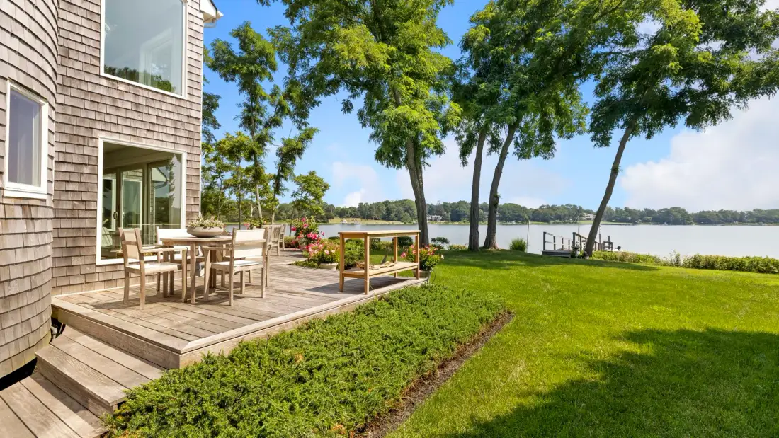 A lakeside house opens onto a wooden deck, lawn, and leafy trees.