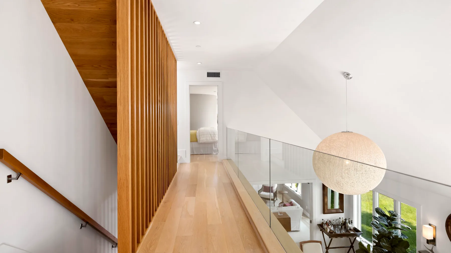 A bright upstairs hallway overlooks a living area, with a glass railing and wood flooring.