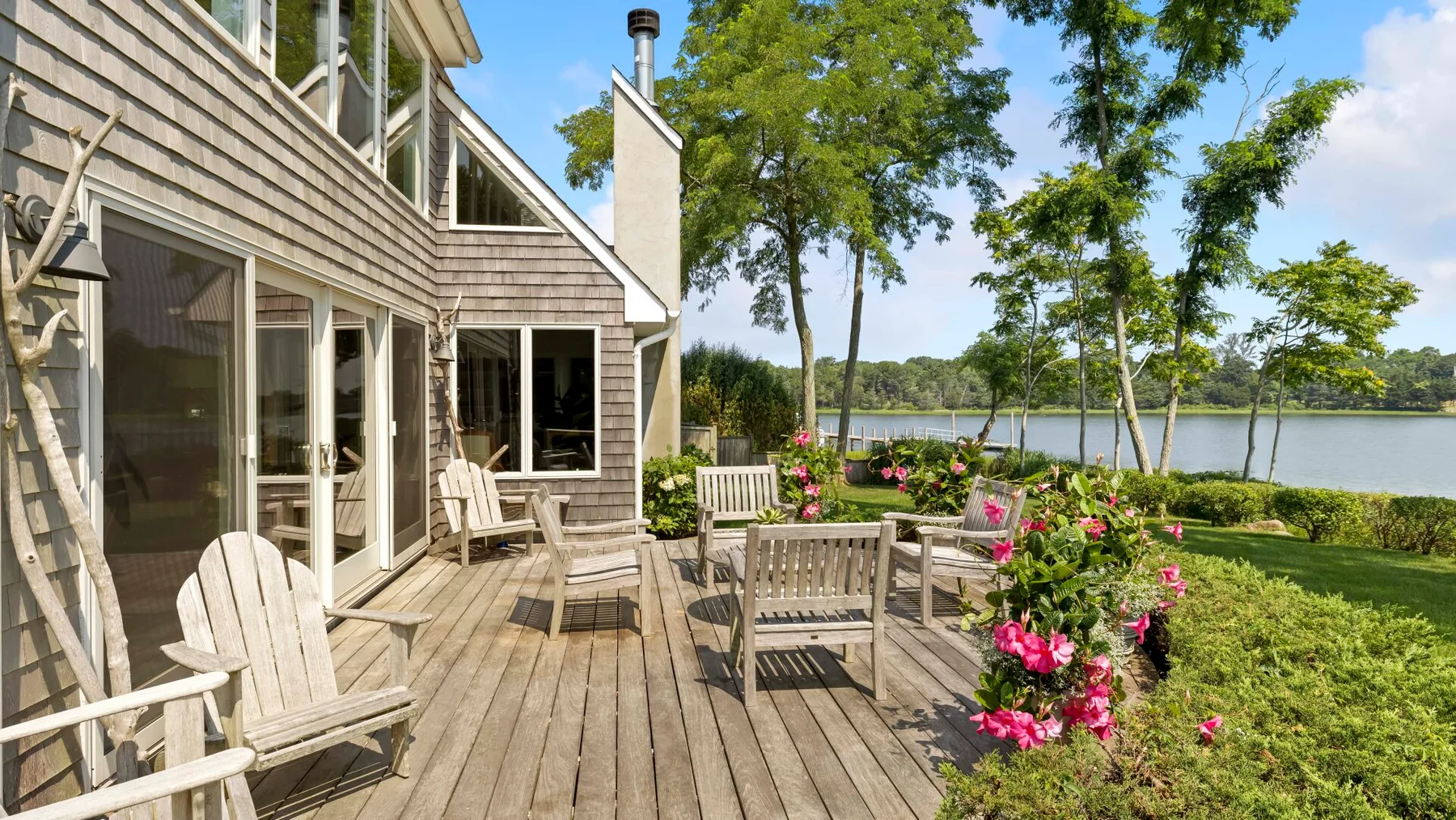 A wooden deck beside a house overlooks a flower garden, lawn, and lake.