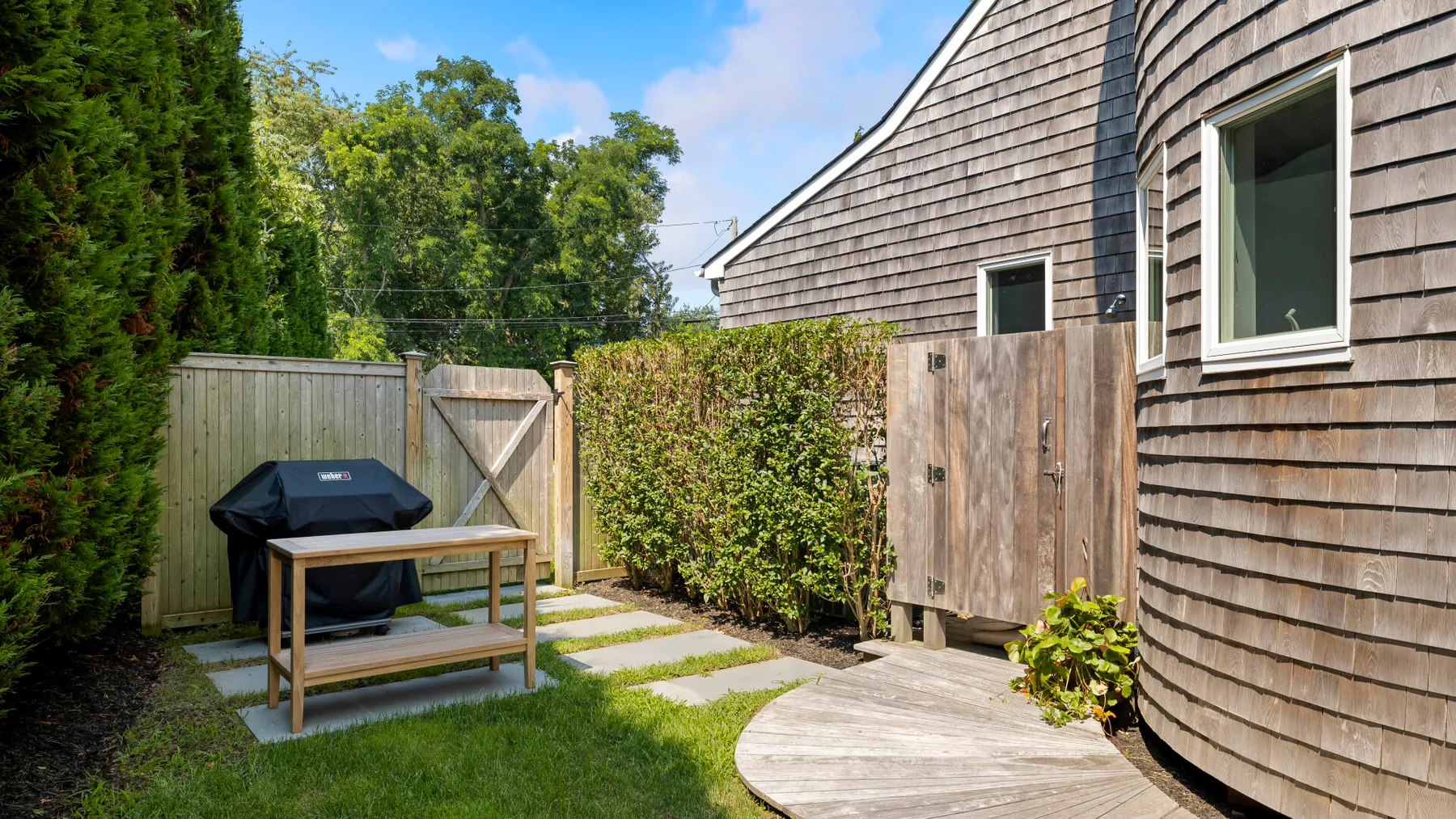 A small outdoor patio beside a shingled building, with a grill and garden path.