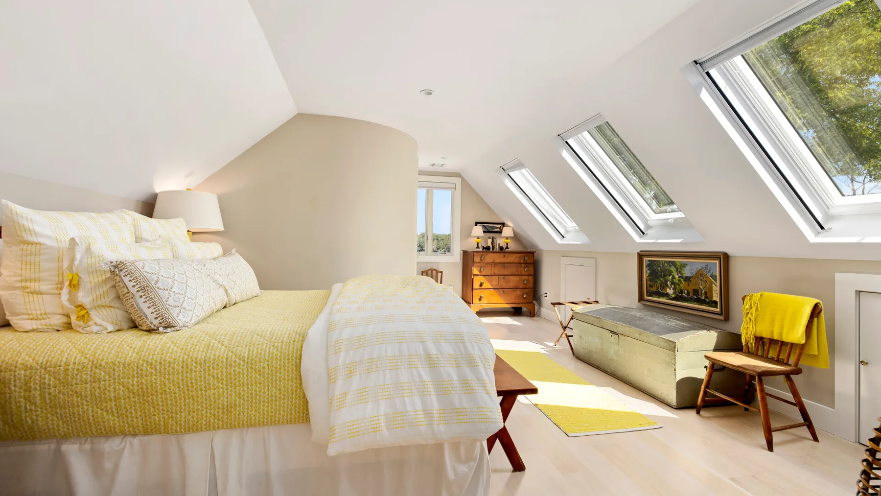 A bright attic bedroom with a bed, low seating, and sloped windows.