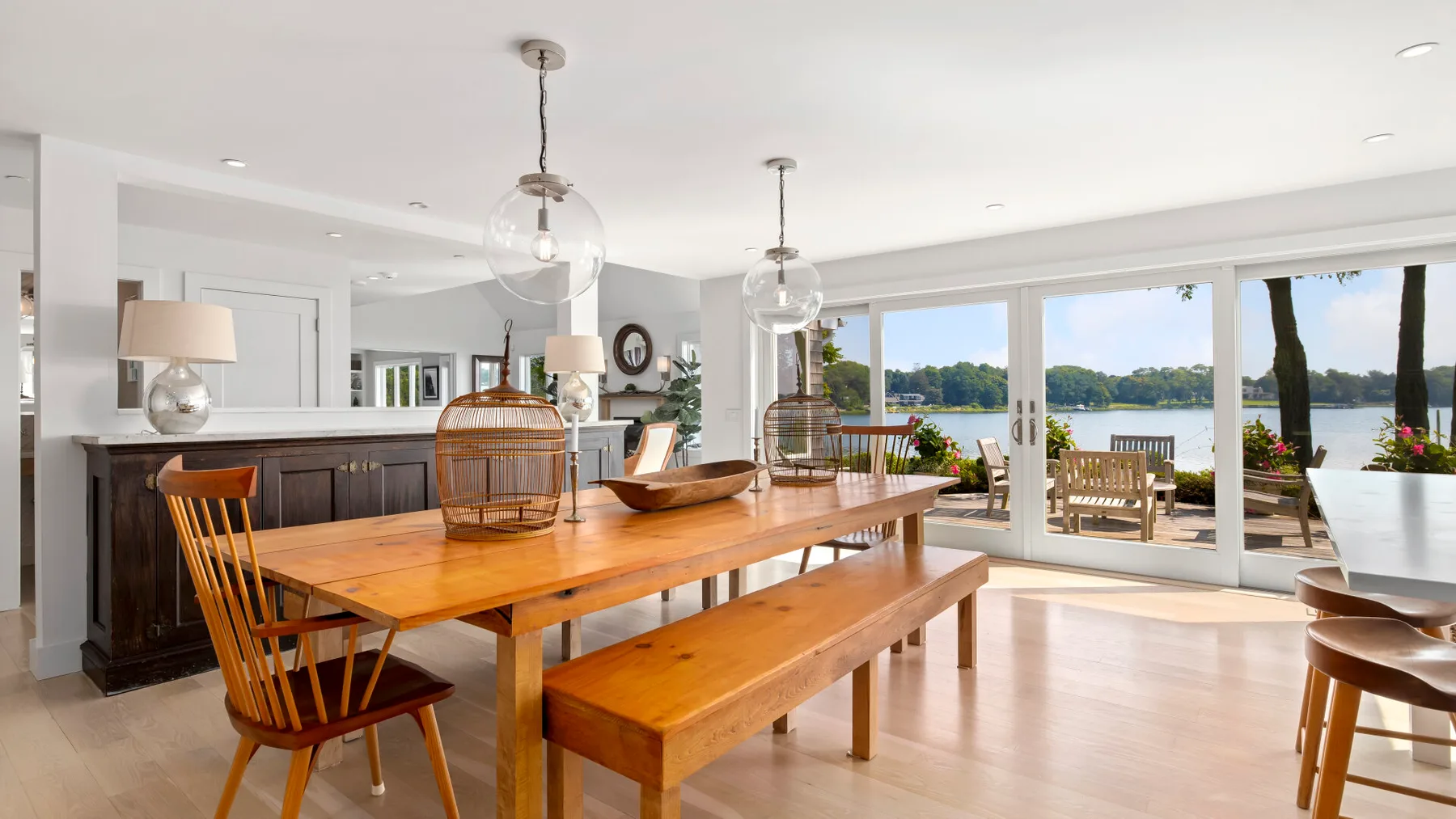A bright dining area has a wooden table and bench beside large windows overlooking the water.