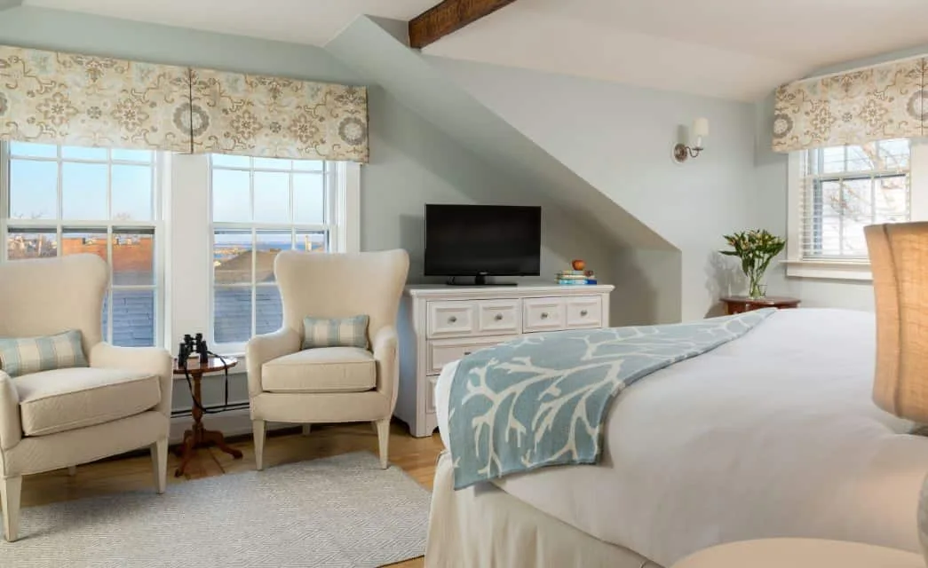 Habitación con piso de madera, paredes azul claro, cómoda blanca, ropa de cama blanca y sillas tapizadas en color crema