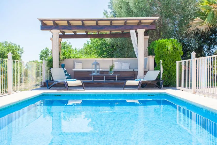 Una piscina está frente a una zona de descanso a la sombra bajo una pérgola.