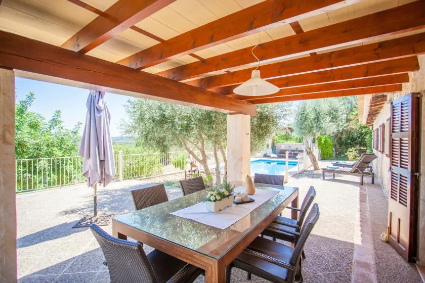 Una zona de comedor en un patio cubierto da a una piscina y vegetación.