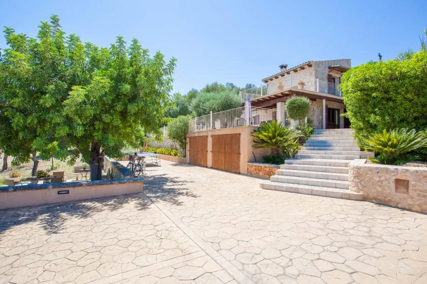 Un patio pavimentado conduce a una casa con amplios escalones y árboles frondosos.