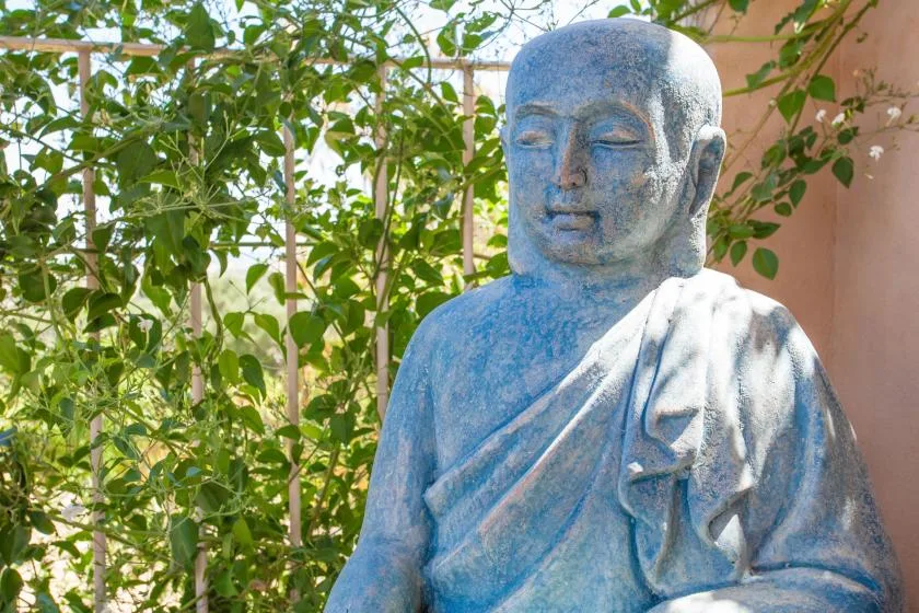 Una estatua azul de una figura sentada está entre plantas frondosas.