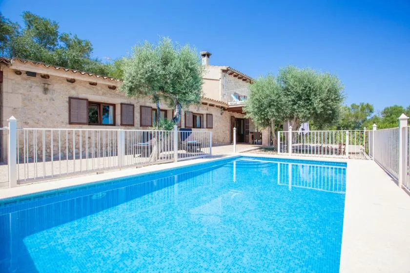 Una piscina exterior luminosa está junto a una casa de piedra y árboles frondosos.