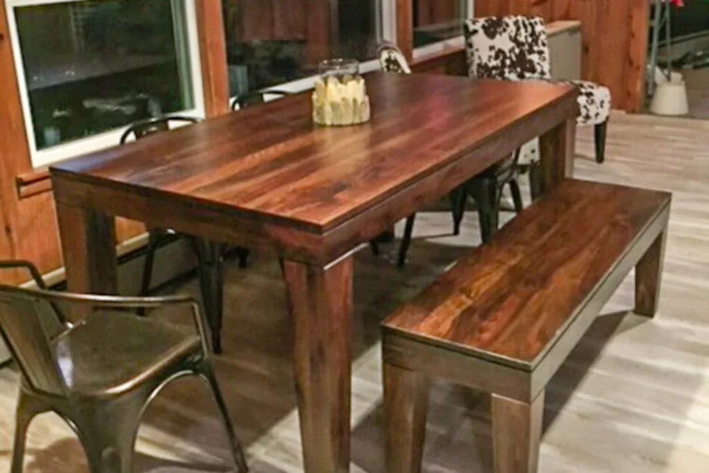 Una mesa de comedor de madera con un banco y sillas está junto a grandes ventanas.