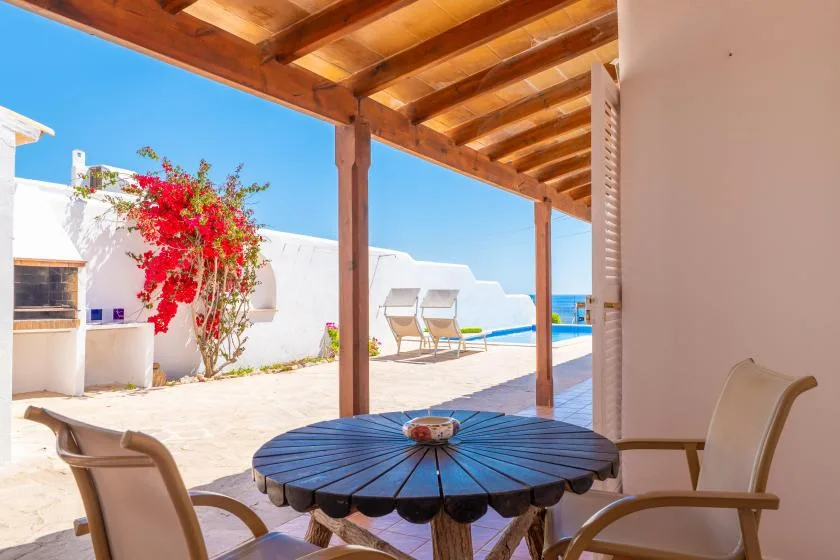 Un patio a la sombra da a una piscina, con una mesa redonda y asientos.