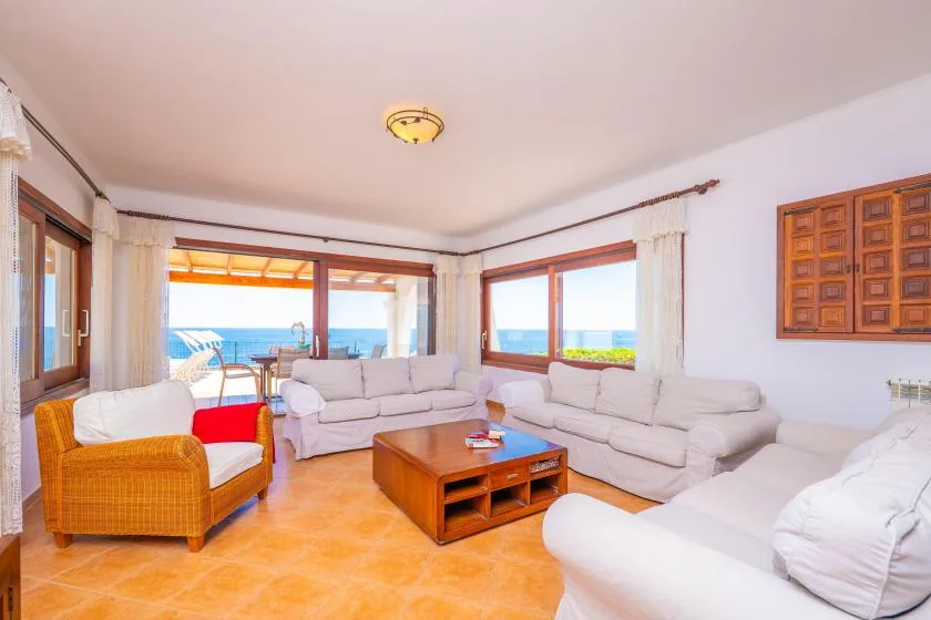 Una sala luminosa con asientos blancos, una mesa de centro de madera y vistas al mar por las ventanas.