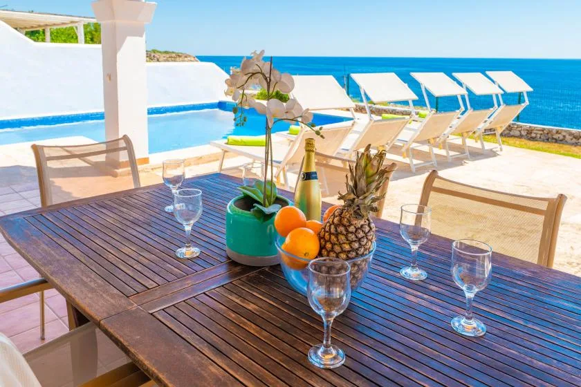 Una mesa de comedor al aire libre con fruta y bebidas da a una piscina y al mar.