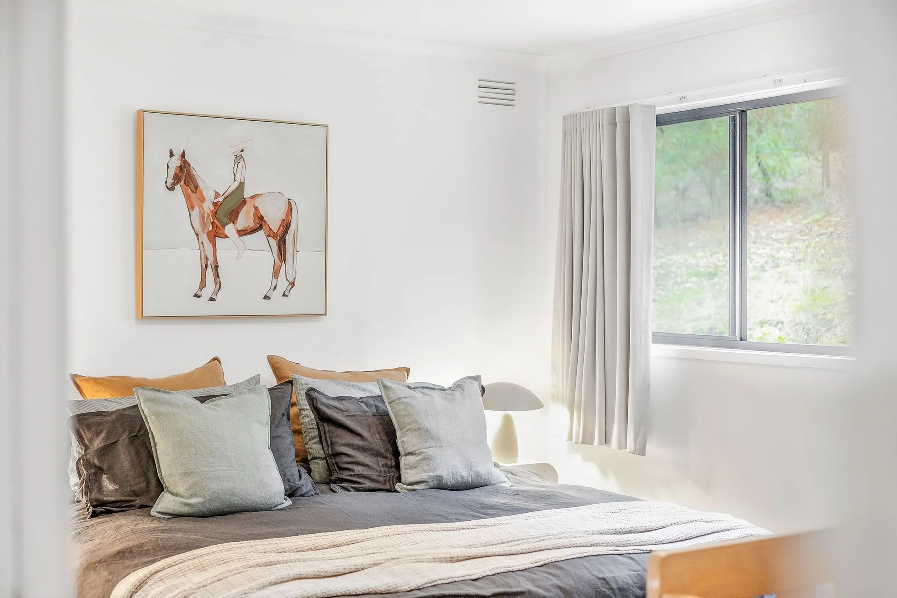 A bright bedroom with a bed, horse artwork, and a large window.
