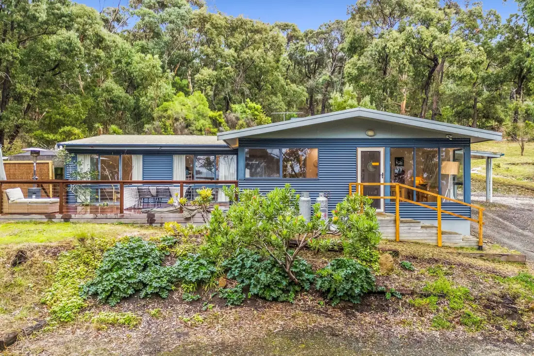 A blue-sided house stands among trees, with a deck and ramp in front.