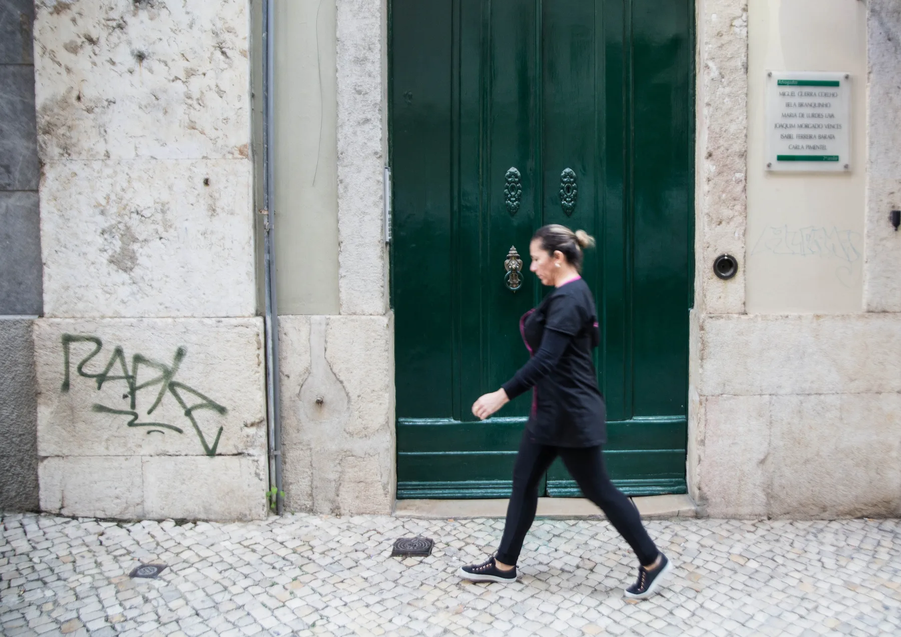 A woman walks past a dark green doorway on a cobblestone street.