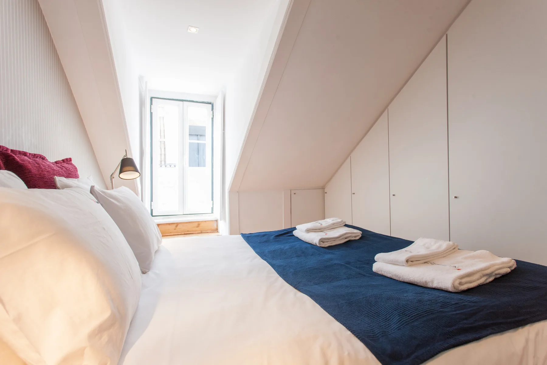 A bright attic bedroom with white bedding and folded towels on a blue bedspread.
