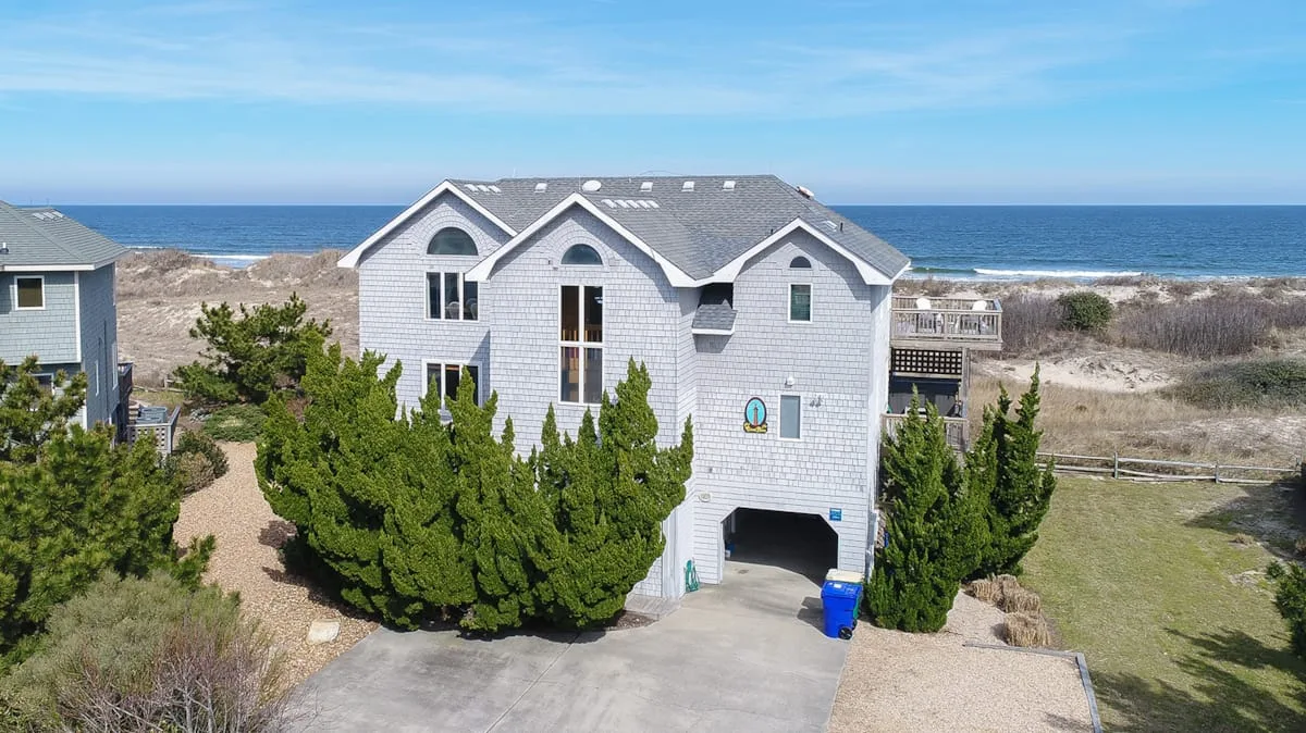 A coastal house stands beside sandy dunes, with the ocean visible behind it.