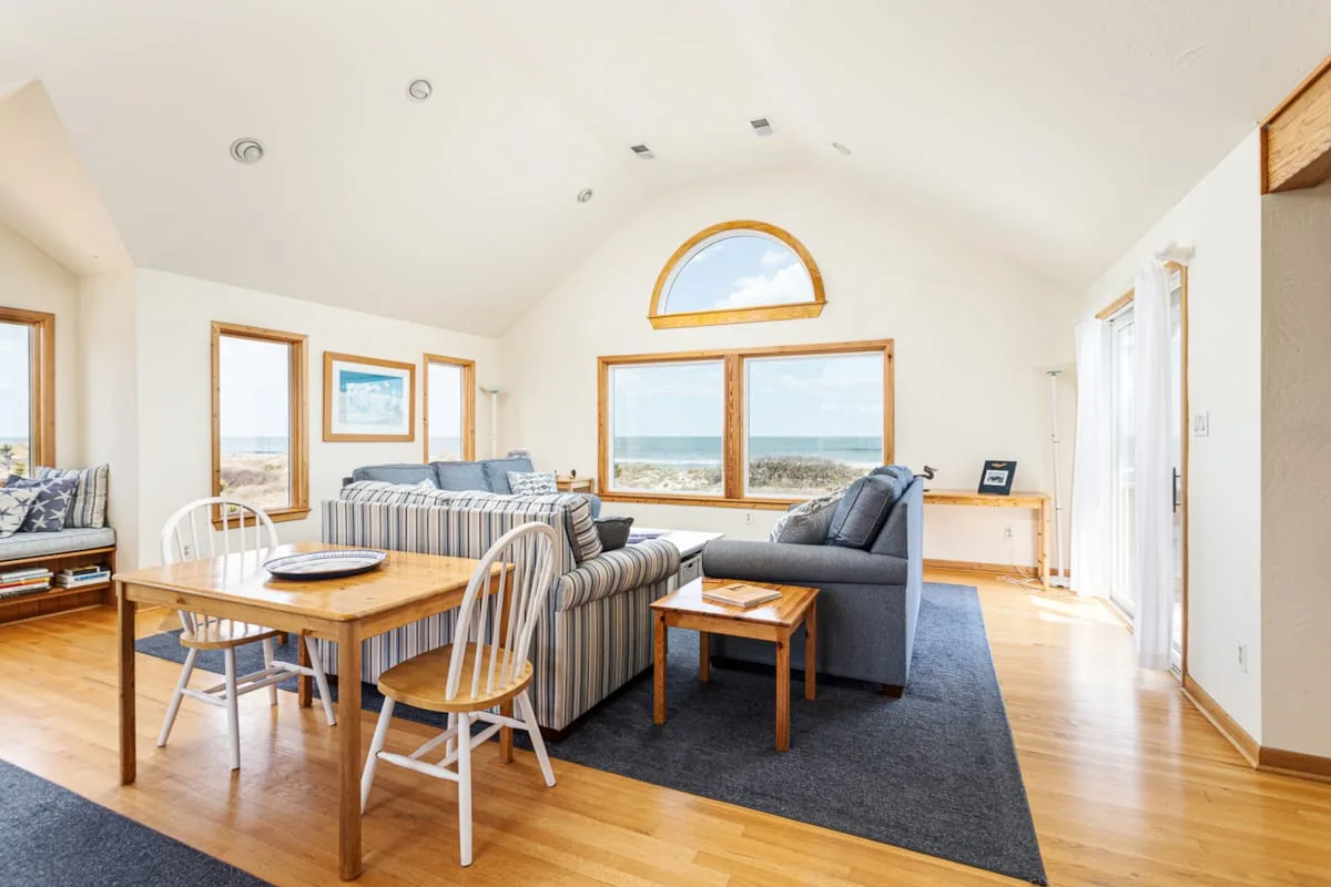 A bright living and dining area with seating beside large ocean-facing windows.