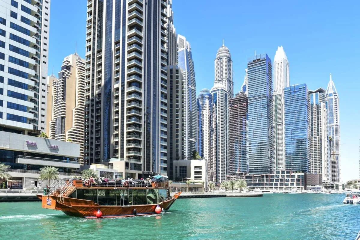 Modern high-rise buildings line a waterfront canal, with a wooden boat in the foreground.