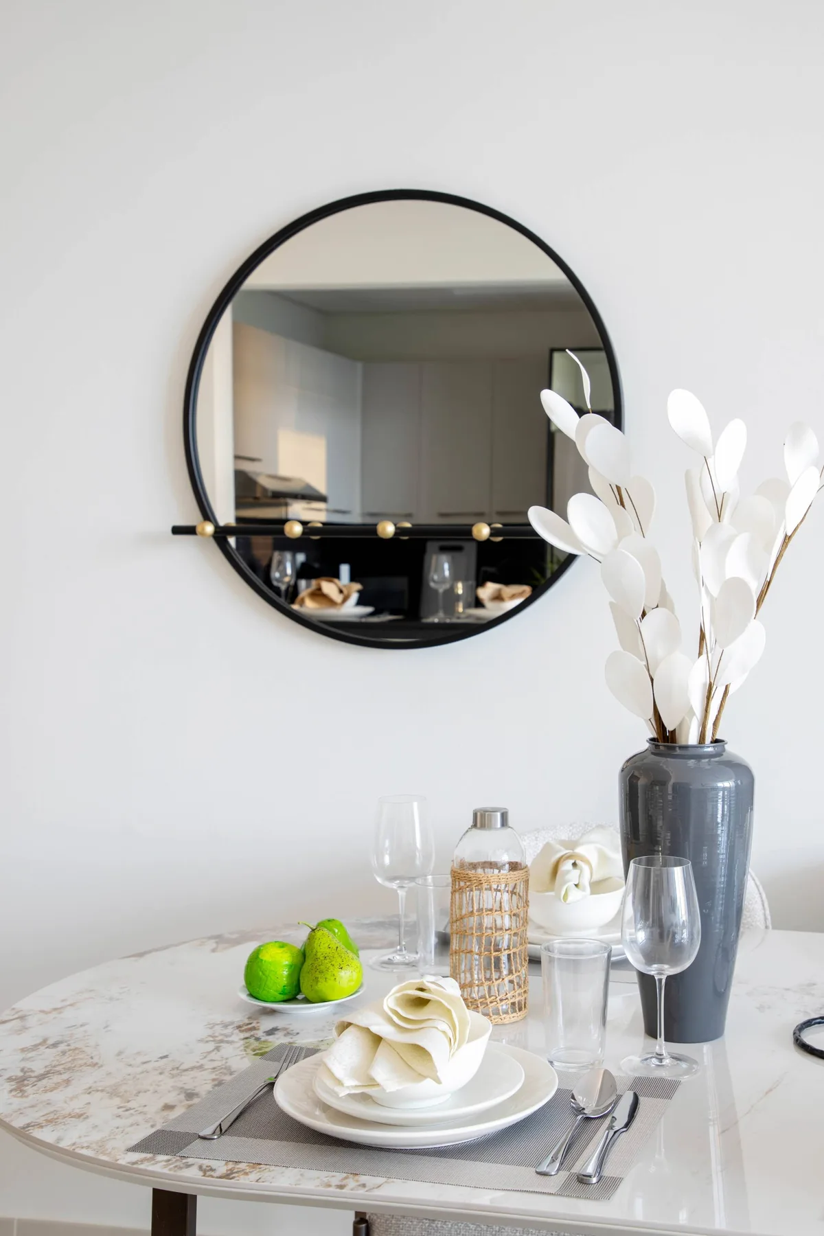 A dining table is set with plates and glassware beneath a round wall mirror.