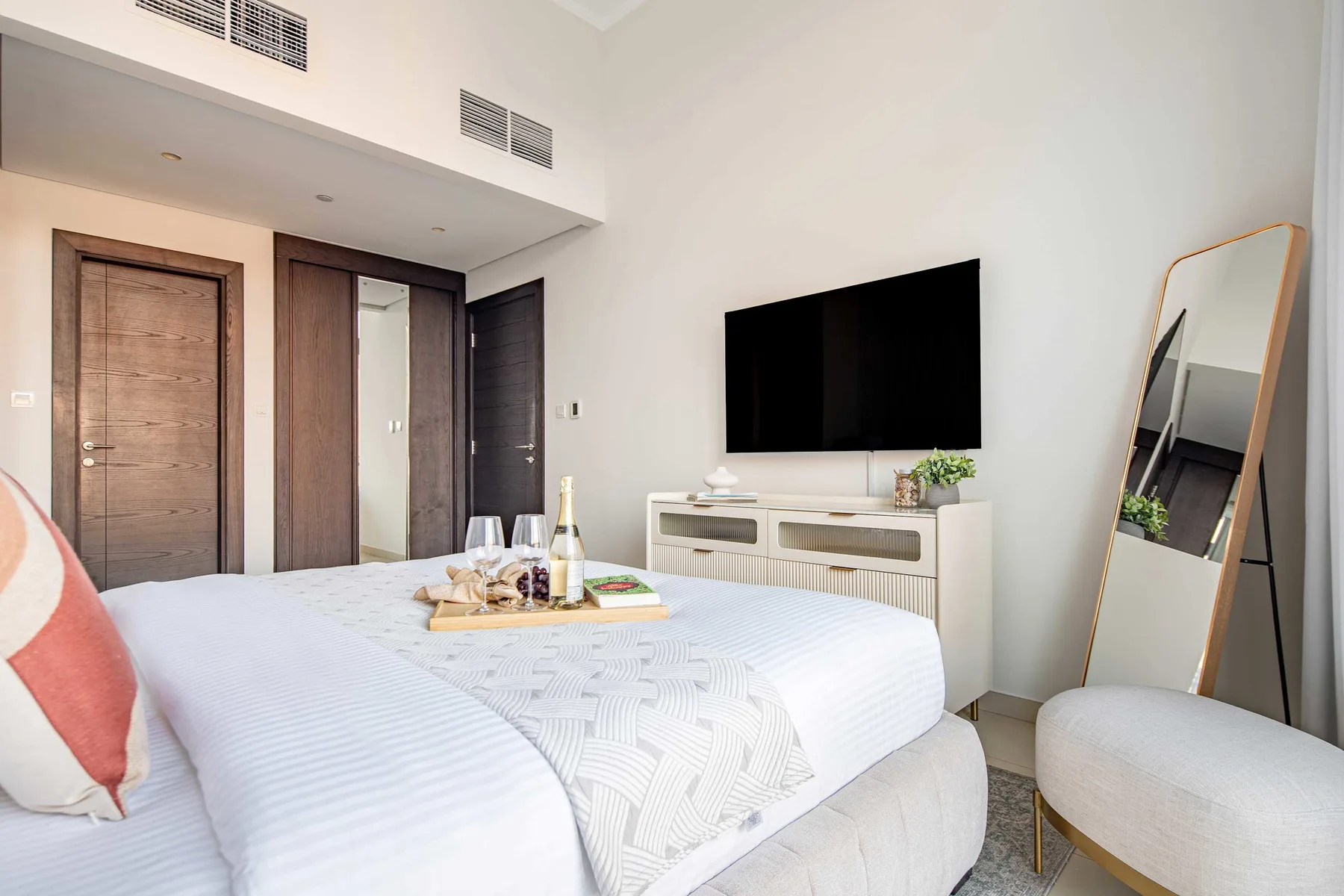 A bright bedroom with a neatly made bed, wall-mounted television, and full-length mirror.