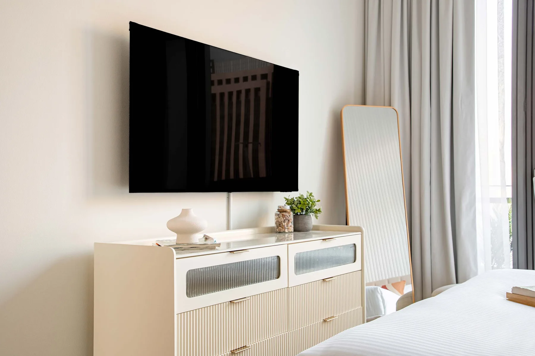A bright bedroom with a television mounted above a dresser beside a curtained window.