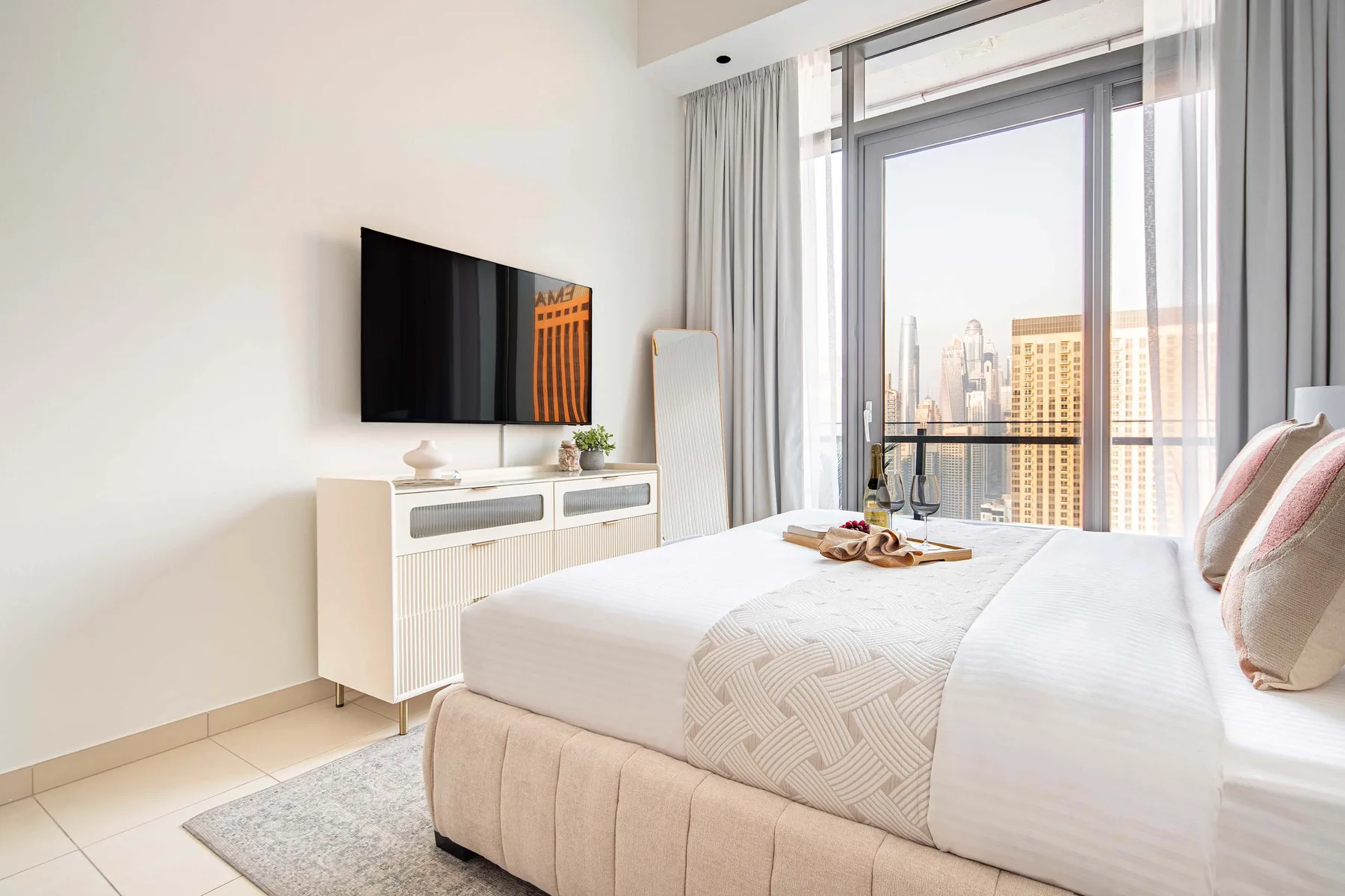 A bright bedroom with a bed beside a large window overlooking city buildings.