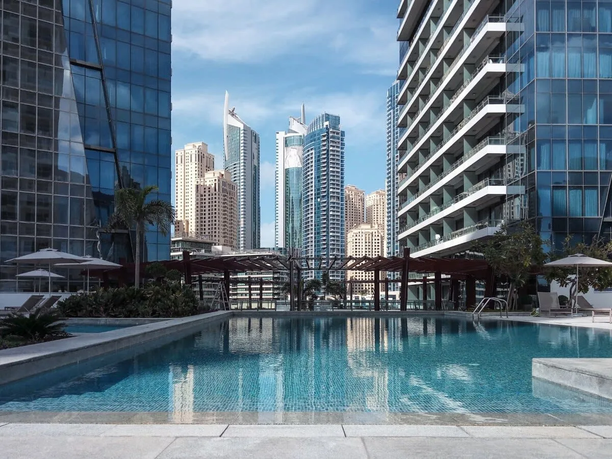 An outdoor swimming pool sits between modern high-rise buildings under a blue sky.