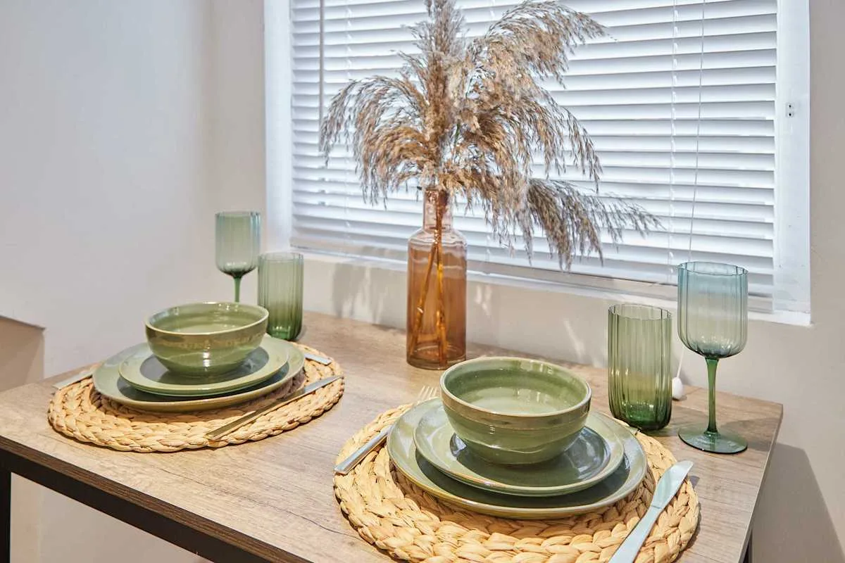 A dining table is set with green dishes and dried flowers by a window.
