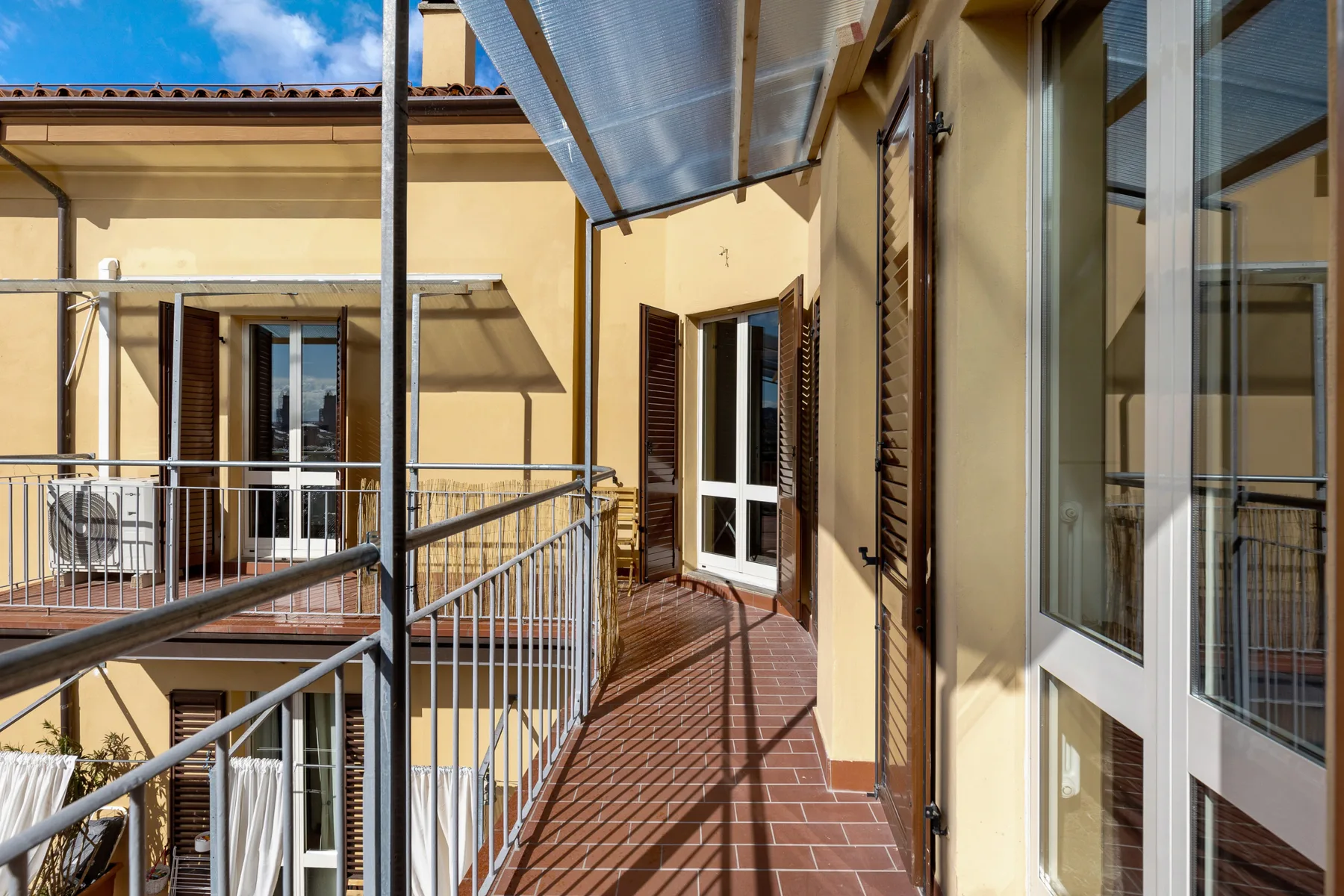 A narrow outdoor balcony walkway with metal railings and shuttered glass doors.