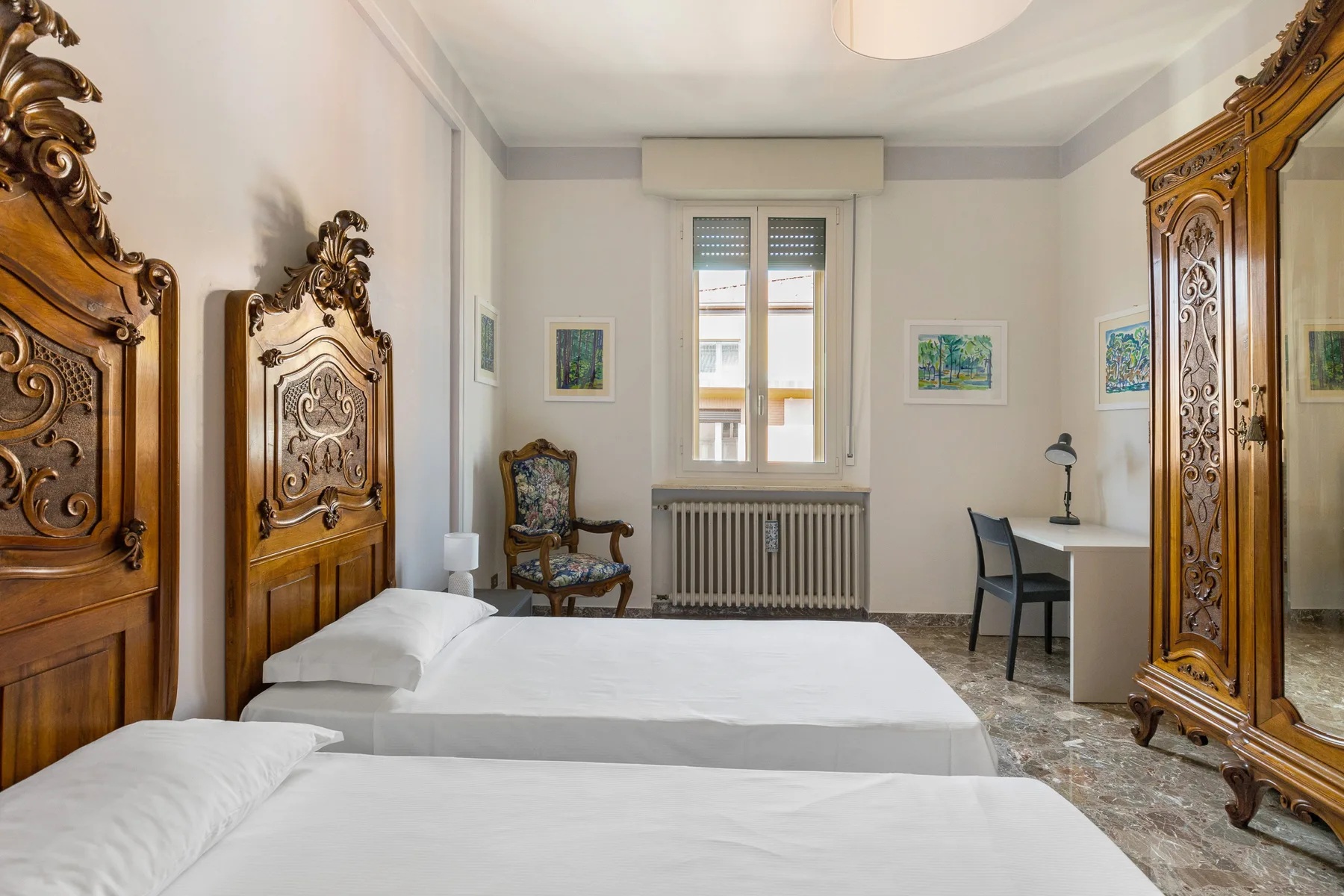 A bedroom with two beds, ornate wooden furnishings, and a desk by the window.