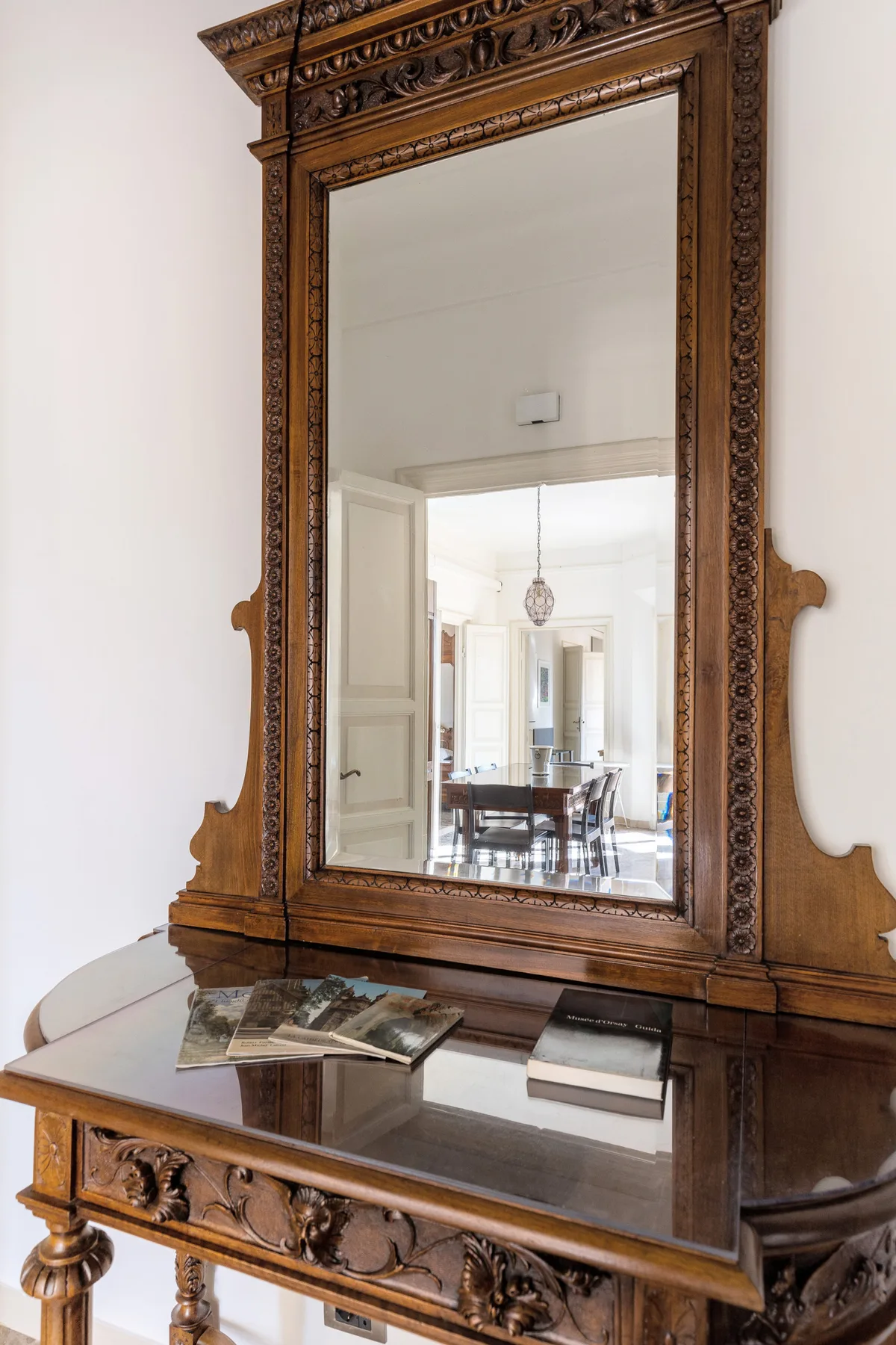 An ornate wooden mirror hangs above a carved console table in a bright interior.