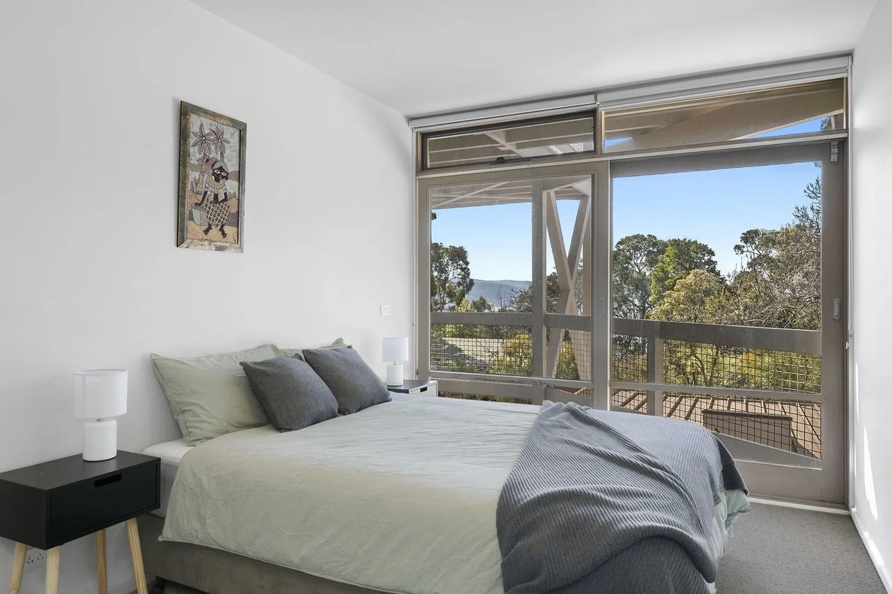A bright bedroom with a bed beside large windows overlooking trees.
